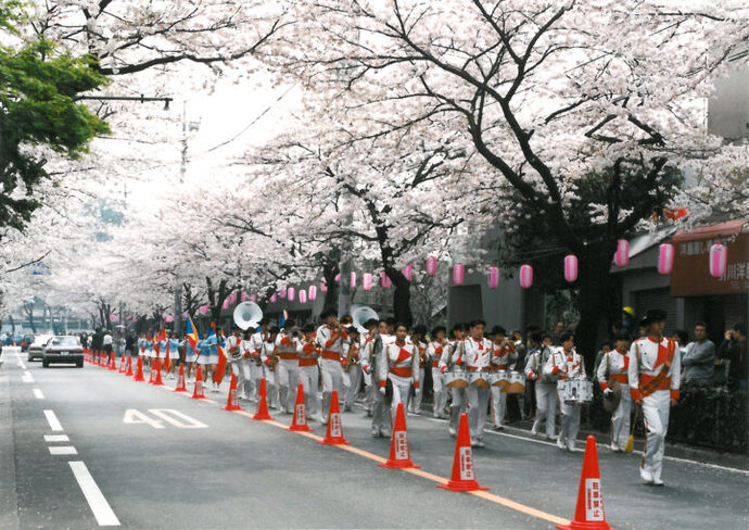 写真：桜の木の下をマーチングバンドが行進する様子